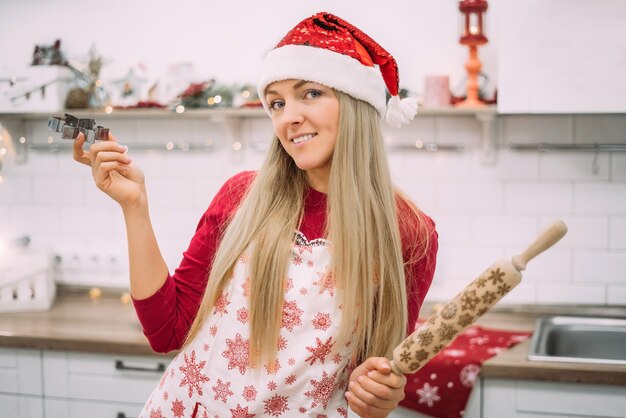 A young woman with blond hair stands in the kitchen wearing an apron and a gingerbread cookie pan in a santa hat