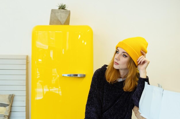 Photo young woman with blond hair in a yellow knitting hat is sitting in a cafe near to yellow fridge