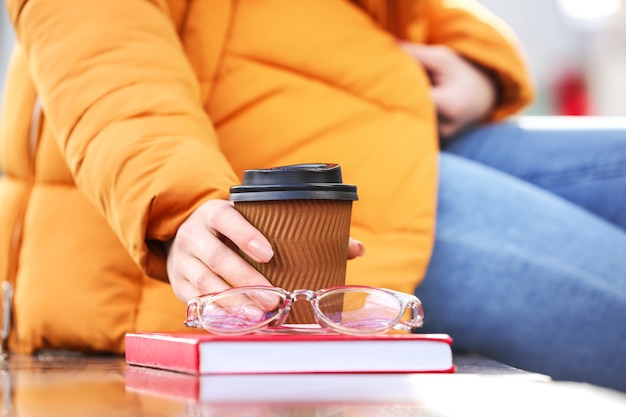 Young woman with book, cup of coffee and eyeglasses sitting on bench