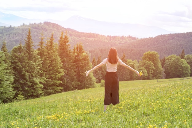 Young woman with a bouquet of flowers and wide open arms standing in the meadow. Forest and mountains in the background. Back view