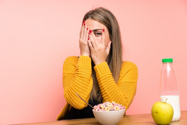 Young woman with bowl of cereals covering eyes and looking through fingers