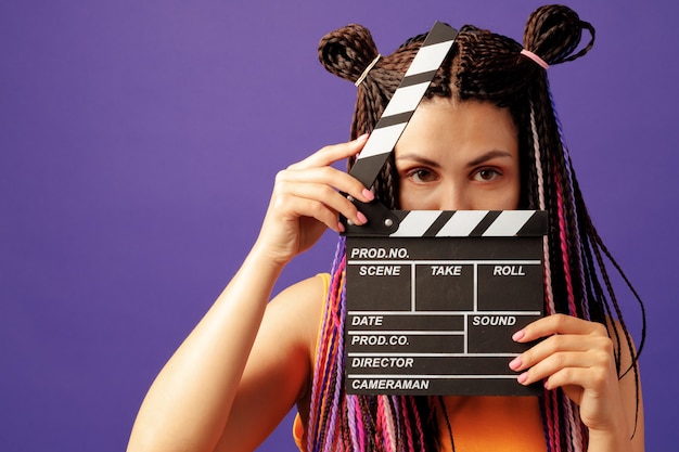 Young woman with braids holding clapper board close-up on purple background