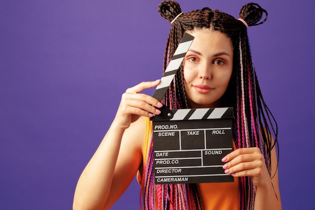 Young woman with braids holding clapper board close-up on purple background