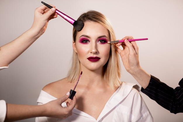 Young woman with bright dark pink makeup posing in studio. Makeup artist's hands correct makeup with a special brush. Professional makeup