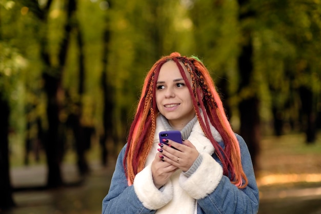 森林公園に立って携帯電話を閲覧する明るいドレッドヘアを持つ若い女性