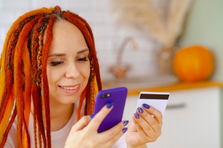 Photo young woman with bright dreadlocks with mobile phone credit card sitting in kitchen happy female browsing smartphone entering card details paying internet purchase concept of online shopping