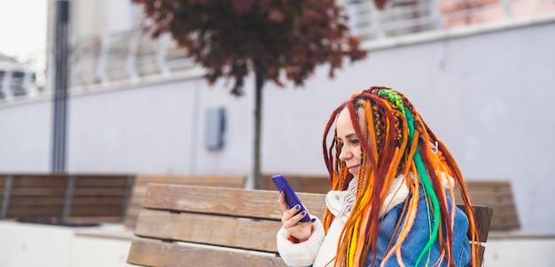 Young woman with bright dreadlocks with mobile phone on walk in park Portrait of positive female in warm clothes browsing smartphone