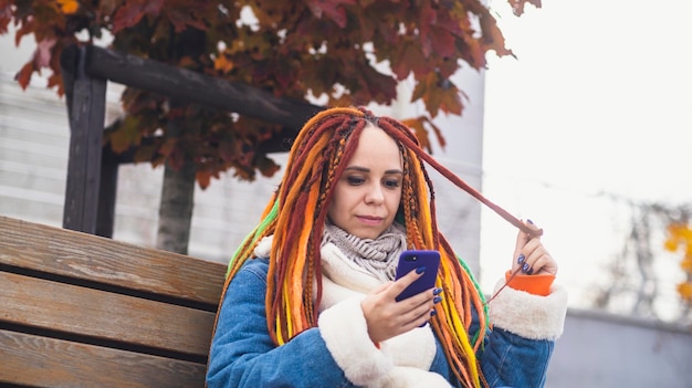 Young woman with bright dreadlocks with mobile phone on walk in park Portrait of positive female in warm clothes browsing smartphone