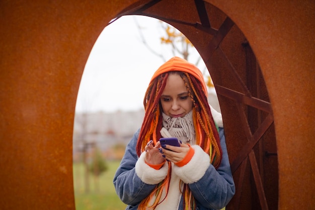 따뜻한 옷을 입은 밝은 헤어스타일을 한 젊은 여성이 눈 덮인 날씨에 공원에 철제 설비에 기대어 휴대폰을 둘러보고 있습니다.