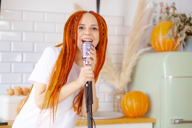 Young woman with bright hairstyle with retro microphone in kitchen Portrait of female singer with dreadlocks singing into microphone at home