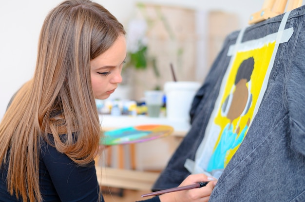 Молодая женщина с кистью, рисующей на джинсах стильную индивидуальную одежду профессионального художника