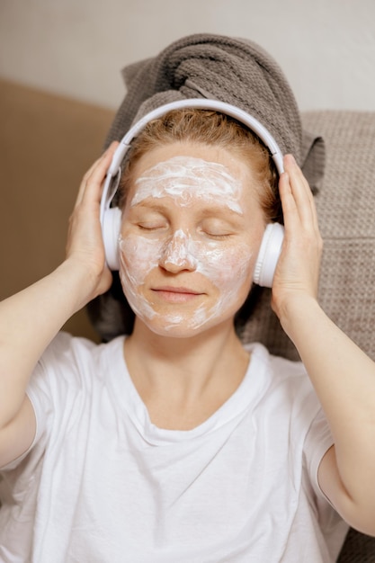 Young woman with casual clothes towel on head and beauty mask on face sitting on couch at home resting and listening music Cosmetic for women skin care Self care time for yourself Relax