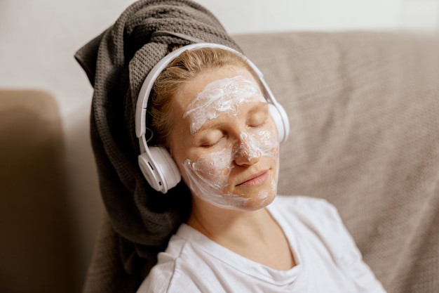 Young woman with casual clothes towel on head and beauty mask on face sitting on couch at home resting and listening music Cosmetic for women skin care Self care time for yourself Relax