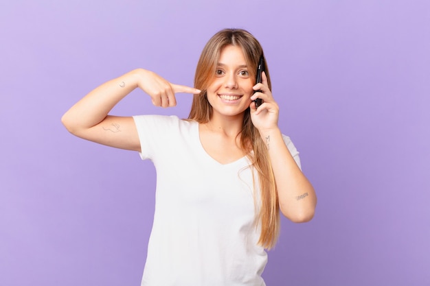 young woman with a cell phone smiling confidently pointing to own broad smile