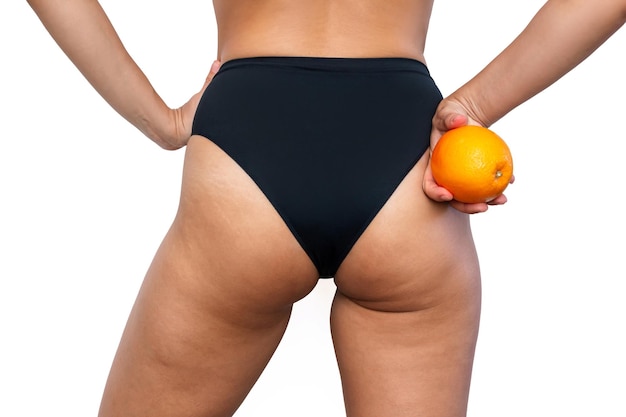 Young woman with cellulite and white stretch marks on thighs stands with her back holding orange