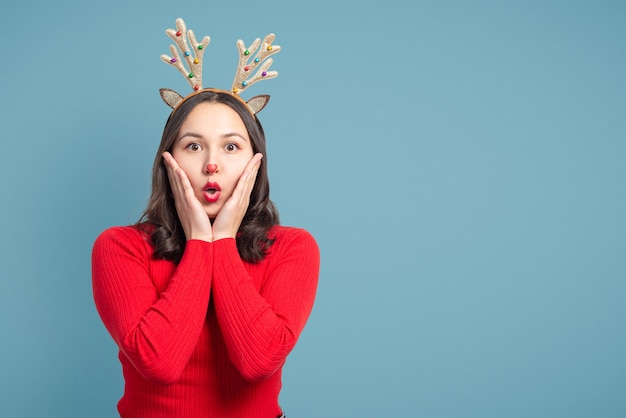 파란색 배경에 빨간 스웨터에 크리스마스 사슴 뿔을 가진 젊은 여자