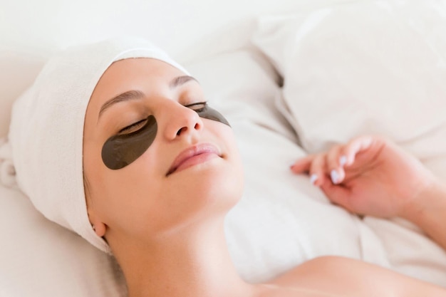 Young woman with closed eyes and black eye patches is lying and relaxing in the bed after having a bath wrapped in towel Beauty treatment and skincare concept