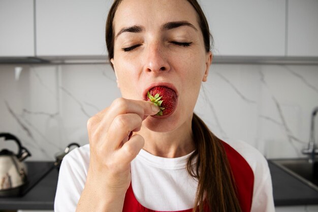 Foto giovane donna con gli occhi chiusi che si diverte a mangiare fragole in cucina