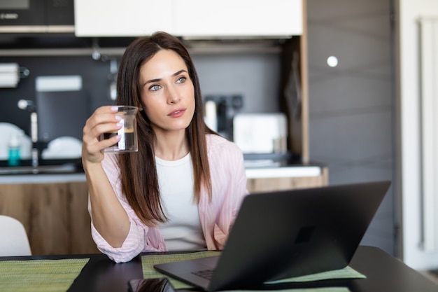 コーヒーカップと自宅の台所でラップトップを持つ若い女性