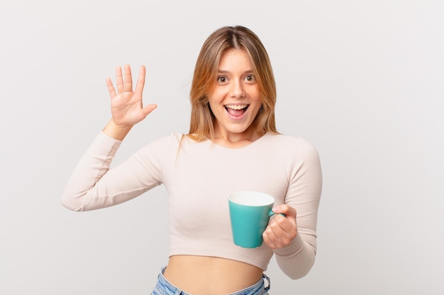 Young woman with a coffee mug smiling happily, waving hand, welcoming and greeting you