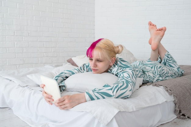 Молодая женщина с цветными розовыми и белыми волосами читает цифровую электронную книгу, лежа дома в постели