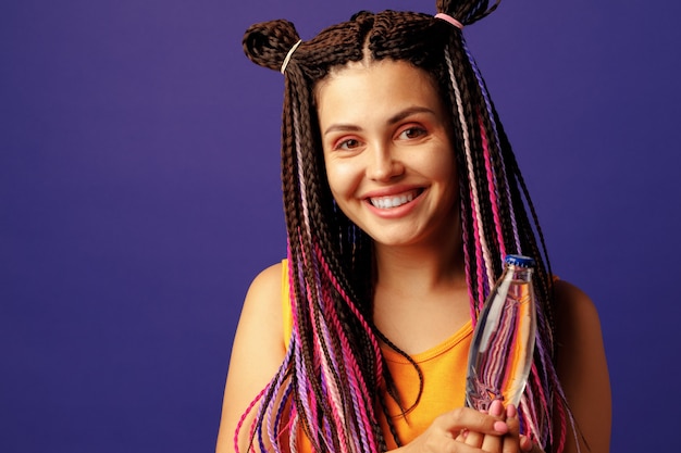 Молодая женщина с красочными афро-косами держит бутылку напитка на фиолетовом фоне