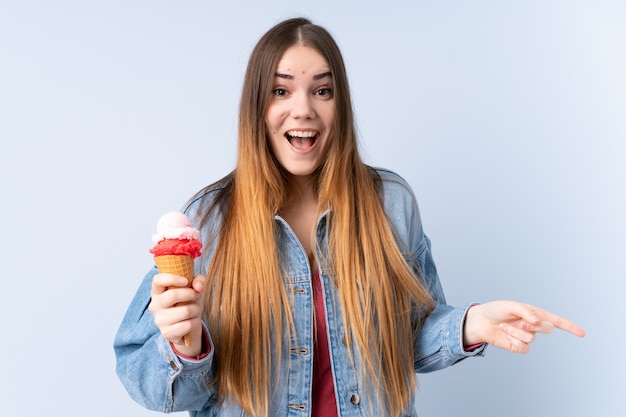 Young woman with a cornet ice cream on blue wall surprised and pointing finger to the side