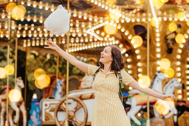 Young woman  with  cotton candy in front of the carrousel with night illumination at the amusement park.