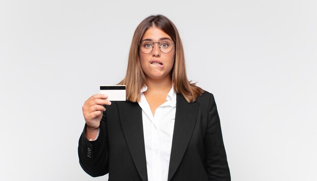 Young woman with a credit card looking puzzled and confused, biting lip with a nervous gesture, not knowing the answer to the problem