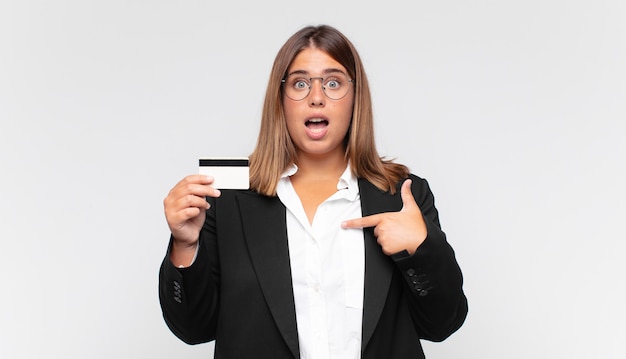 Young woman with a credit card looking shocked and surprised with mouth wide open, pointing to self