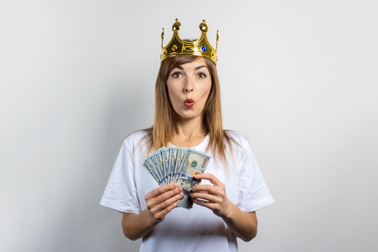 Photo 彼女の頭には王冠と驚いた顔を持つ若い女性は明るい背景にお金のスタックを保持しています。