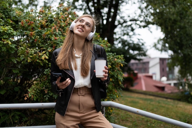 リラックスしながらヘッドフォンで音楽を聴く携帯電話でコーヒーのカップを持つ若い女性