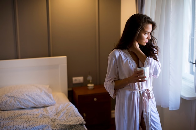 Young woman with cup of coffee or tea in neglige standing by the window