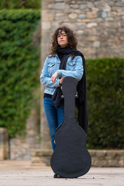 Young woman with curly hair and glasses posing with a guitar case in the street on a cloudy day