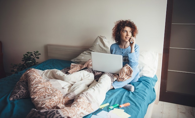 Young woman with curly hair is talking on phone and using a laptop in bed early morning wearing a blue pajama and doing homework