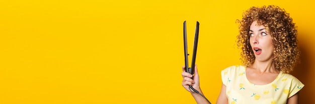 Young woman with curly hair looks in surprise at the hair straightener on a yellow background