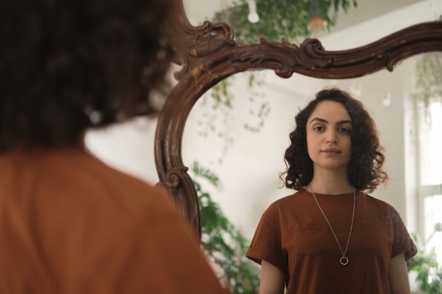 Молодая женщина с вьющимися волосами стоит перед зеркалом и смотрит на себя