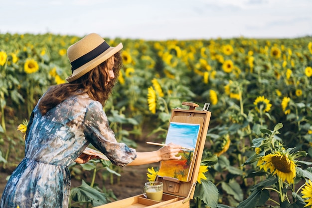 A young woman with curly hair and wearing a hat is painting in nature. A woman stands in a sunflower field on a beautiful day