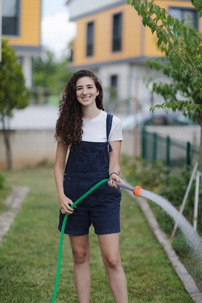 Photo young woman with curly hair wearing overall watering plants in country house backyardm smiling looking to camera garden care ecofriendly lifestyle suburban life concept
