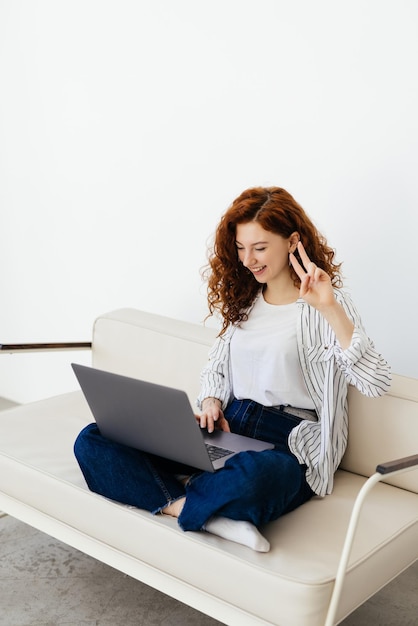 Молодая женщина с вьющимися рыжими волосами лежит на диване и участвует в видеоконференции на внештатной и удаленной работе на ноутбуке