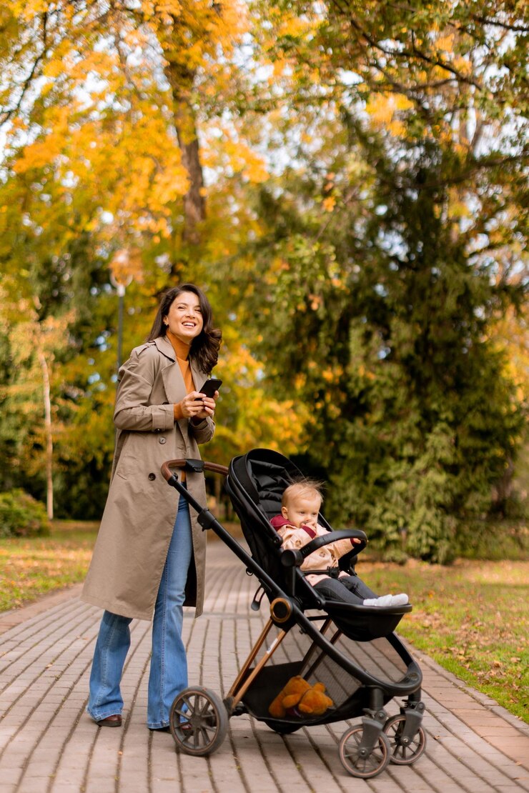Photo young woman with cute baby girl in baby stroller using mobile phone at the autumn park