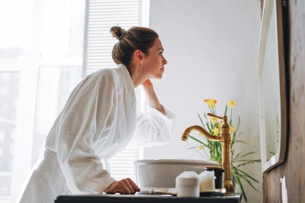 Photo young woman with dark long hair in white bathrobe near mirror in bathroom at home