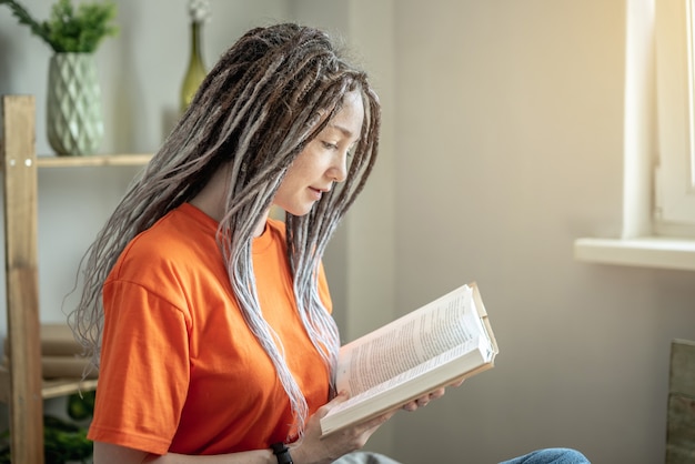 Photo young woman with dreadlocks and bright clothes is sitting and reading a book at home