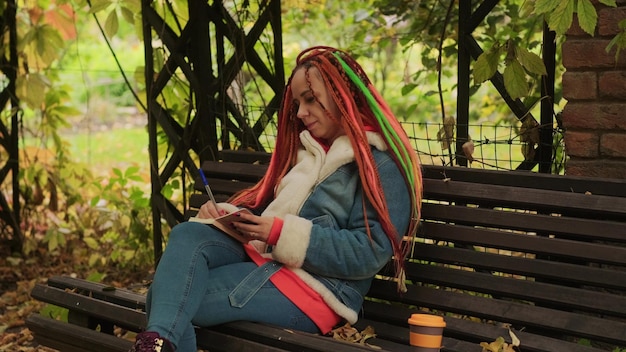 Young woman with dreadlocks drawing writing in notebook sitting on bench under archway with flowering vegetation