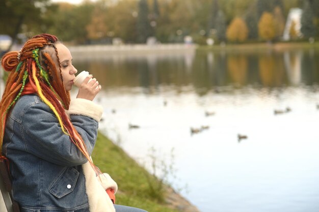 Young woman with dreadlocks in warm clothes sits near lake drinking coffee in park