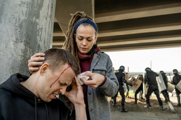 Young woman with dreads putting bandage to head of man sufferred in struggle of rebels and riot police, woman helping to stop blood to victim