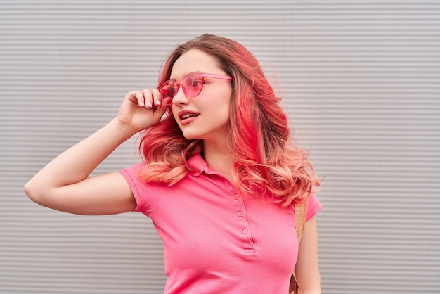 Giovane donna con acconciatura tinta rosa e occhiali da sole sorridente sullo sfondo grigio