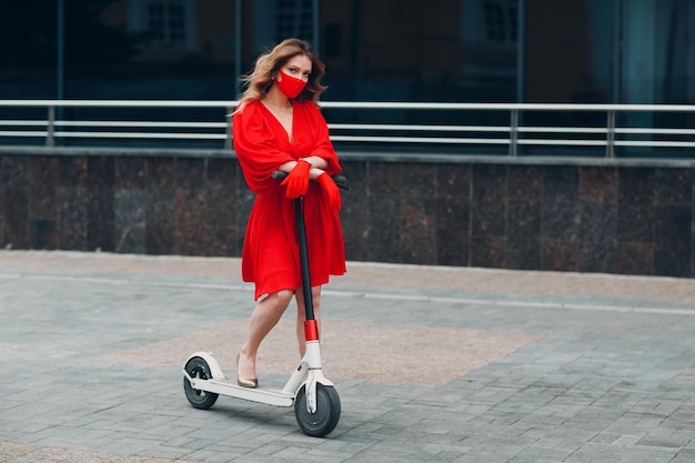 Young woman with electric scooter in red dress and gloves with\
face mask at the city new normal fashion and coronavirus covid\
protection concept