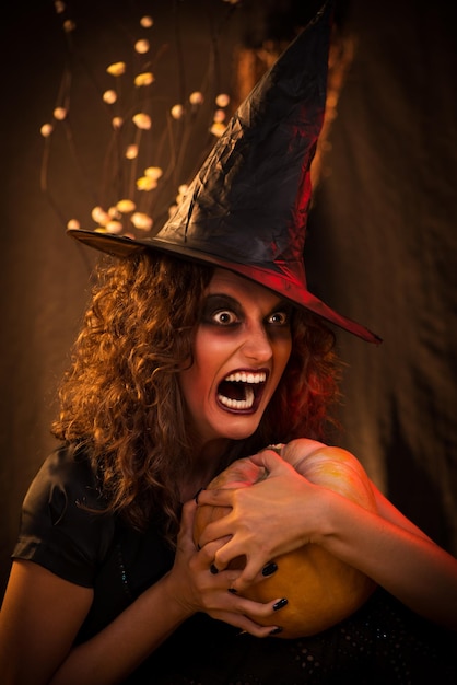 Giovane donna con la faccia malvagia vestita come una strega. indossa abiti scuri e tiene in mano una zucca.