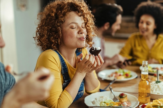 Молодая женщина с закрытыми глазами наслаждается вкусом еды во время еды с друзьями за обеденным столом.